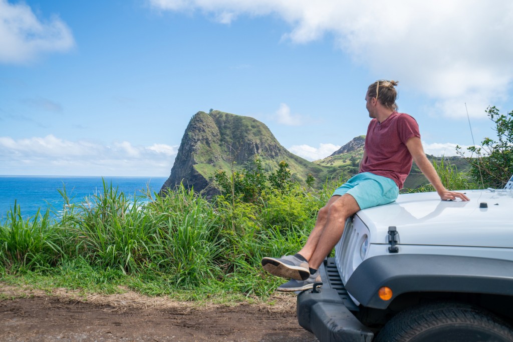Young Man On Road Trip Looking At Coastline From Vehicle Hood