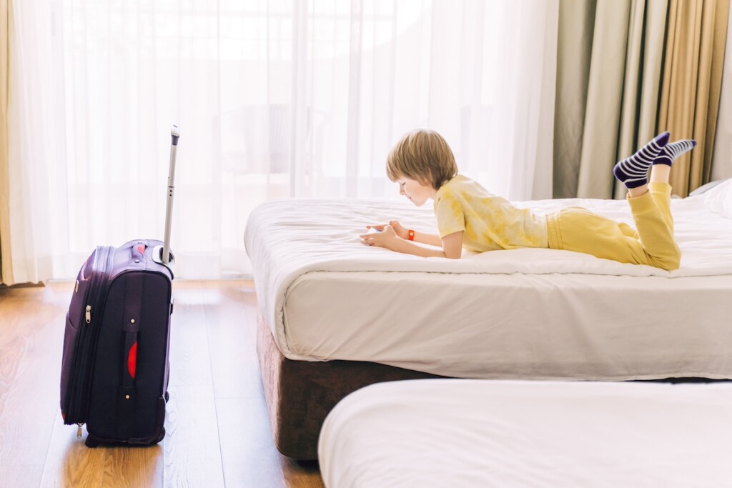 6 Years Boy Have A Fun And Play The Phone In Hotel Room. Suitcase On The Floor Near Bed.